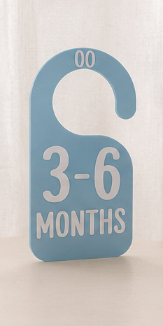A light blue baby door hanger with white text reading '3-6 MONTHS' and '00' at the top, shown standing against a soft beige curtain background.